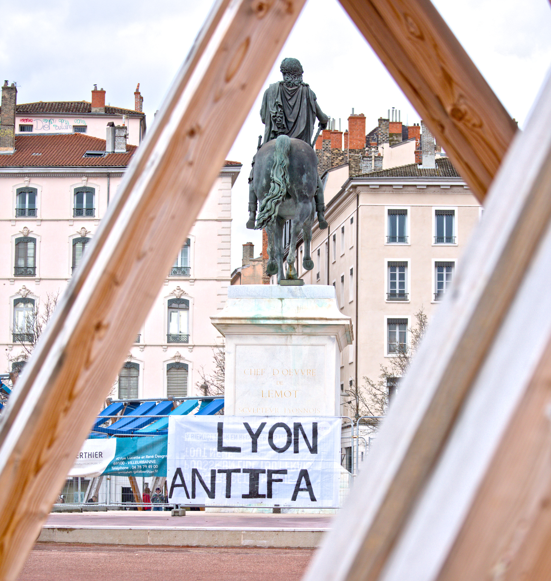 Banderole « Lyon antifa » au pied d’une statue équestre. Un peu partout à l’avant-plan, des bouts d’une grande structure en bois.